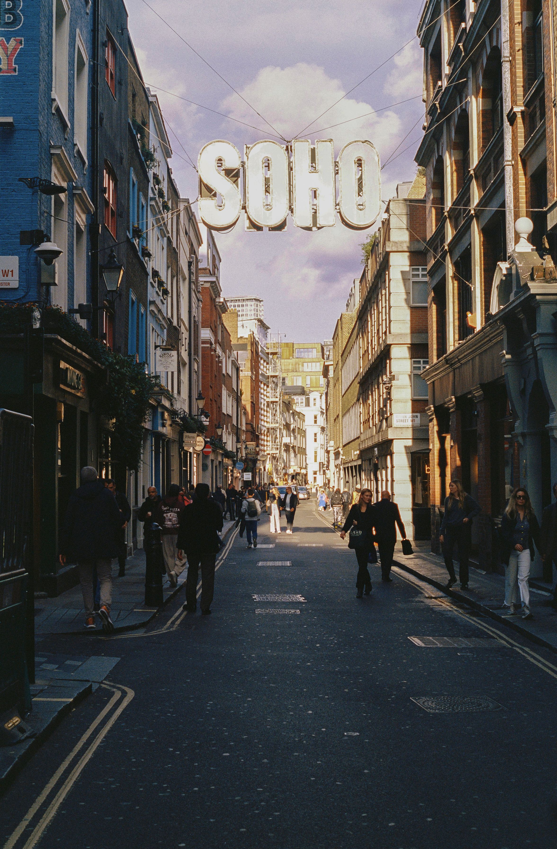 A busy Soho street in London with pedestrians, historic façades and the Soho sign overhead.