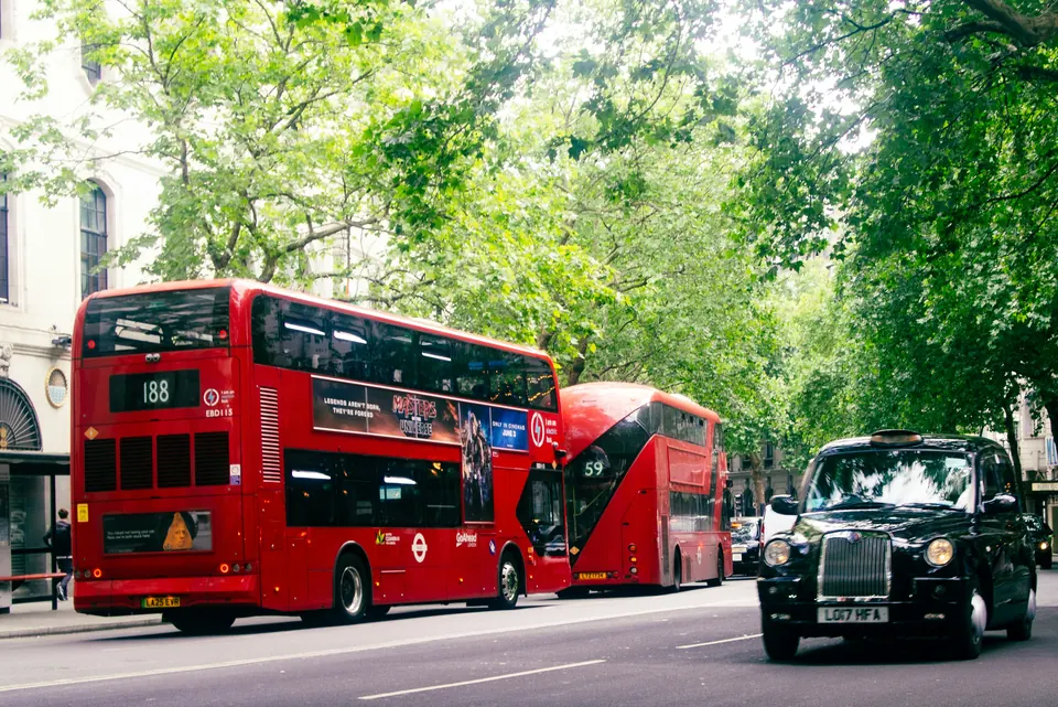 Red London buses and a black taxi travelling along a tree-lined London street.